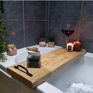 May include: A wooden bath tray with a black handle, a glass jar of bath salts, two candles, and a glass of red wine. The tray is sitting on the edge of a bathtub filled with bubbles.