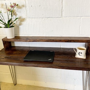 May include: A dark brown wooden desk with a shelf above the desktop. The desk has hairpin legs and a laptop computer on the desktop. A white mug with the letter "e" on it is on the shelf.