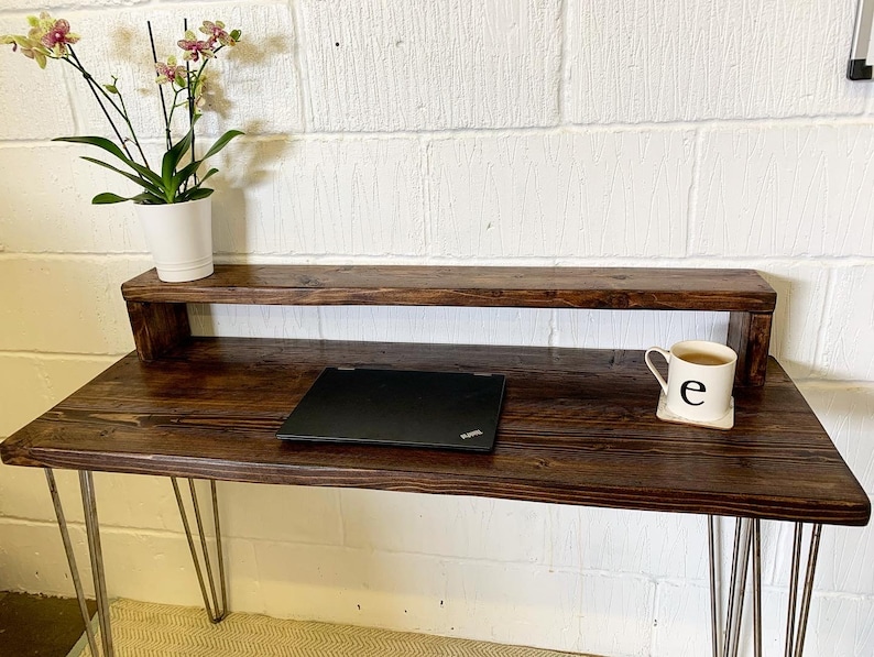 May include: A dark brown wooden desk with a shelf above the desktop. The desk has hairpin legs and a laptop computer on the desktop. A white mug with the letter "e" on it is on the shelf.