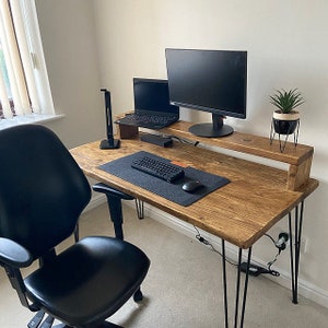 May include: A wooden desk with a black metal frame and hairpin legs. The desk has a shelf for a monitor and a laptop. There is a black keyboard and mouse on the desk. A black office chair is in the foreground.