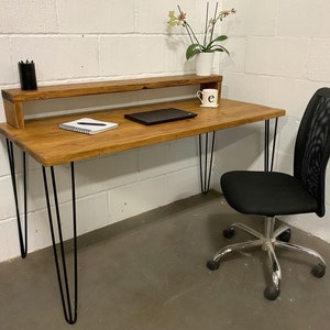 May include: A rustic wooden desk with a black metal shelf and hairpin legs. The desk has a notebook and pen on the surface, a laptop, and a small potted plant. A black mesh office chair with silver wheels is positioned next to the desk.
