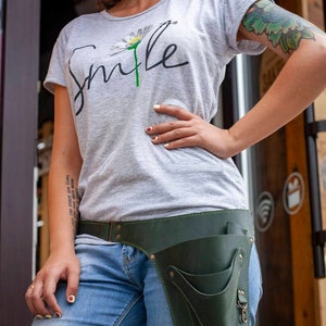 K&ouml;nnte beinhalten: Eine Frau tr&auml;gt ein graues T-Shirt mit dem Wort "Smile" und einem G&auml;nsebl&uuml;mchen-Motiv. Sie tr&auml;gt einen dunkelgr&uuml;nen Leder-Werkzeugg&uuml;rtel mit Taschen und einer Schnalle. Der G&uuml;rtel wird &uuml;ber einer blauen Jeans getragen.