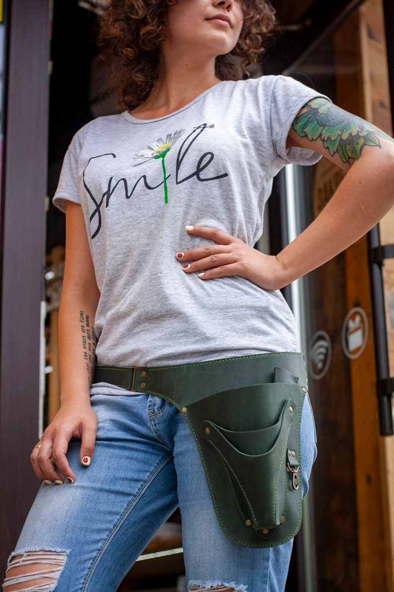 K&ouml;nnte beinhalten: Eine Frau tr&auml;gt ein graues T-Shirt mit dem Wort "Smile" und einem G&auml;nsebl&uuml;mchen-Motiv. Sie tr&auml;gt einen dunkelgr&uuml;nen Leder-Werkzeugg&uuml;rtel mit Taschen und einer Schnalle. Der G&uuml;rtel wird &uuml;ber einer blauen Jeans getragen.