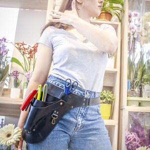 K&ouml;nnte beinhalten: Eine Frau tr&auml;gt ein graues T-Shirt und eine blaue Jeans mit einem schwarzen Leder-Werkzeugg&uuml;rtel mit verschiedenen Werkzeugen, darunter eine Schere und ein Messer. Der G&uuml;rtel wird an der H&uuml;fte getragen und hat eine Metallschnalle.