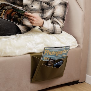 Może przedstawiać: Zielony skórzany organizer do łóżka z dwoma kieszeniami. Organizer jest zawieszony na boku łóżka. W kieszeniach znajdują się czasopisma, telefon i okulary.