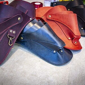 K&ouml;nnte beinhalten: Drei Lederholster in verschiedenen Farben: Burgunderrot, Marineblau und Orange. Jedes Holster hat ein einzigartiges Design mit N&auml;hten und Metallakzenten.