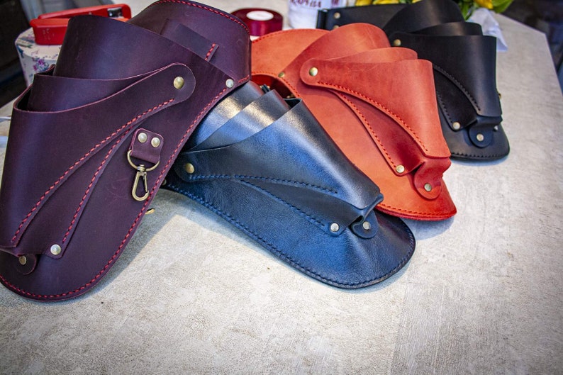 K&ouml;nnte beinhalten: Drei Lederholster in verschiedenen Farben: Burgunderrot, Marineblau und Orange. Jedes Holster hat ein einzigartiges Design mit N&auml;hten und Metallakzenten.