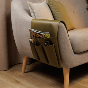 May include: Olive green leather sofa armrest organiser with multiple pockets. A magazine, remote controls, and other items are stored in the pockets. The organiser is attached to a beige sofa with wooden legs. The magazine reads "Photographer".