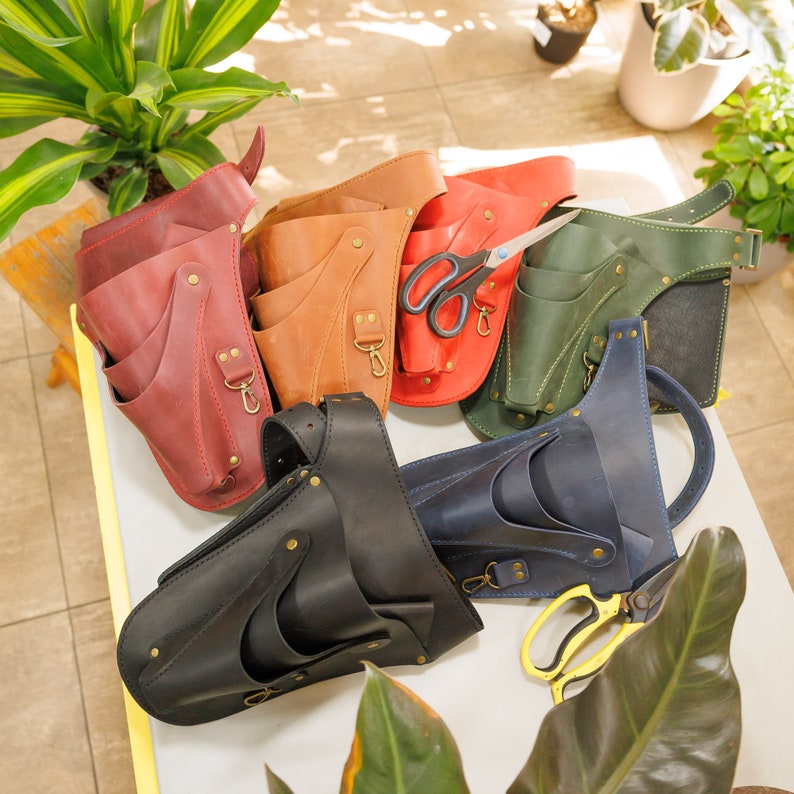 May include: Five leather tool belts with multiple pockets and loops, in various colors including brown, black, green, and red. The belts are arranged on a light brown surface with a pair of scissors and a pair of gardening shears.
