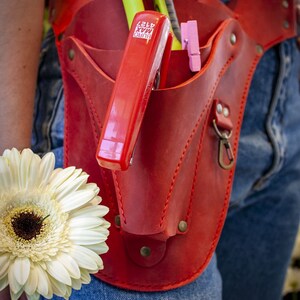 K&ouml;nnte beinhalten: Ein roter Leder-Werkzeugg&uuml;rtel mit einem roten Hefter, einer gr&uuml;nen und silbernen Schere und einem Paar rosa W&auml;scheklammern. Der G&uuml;rtel wird &uuml;ber einer blauen Jeans getragen.
