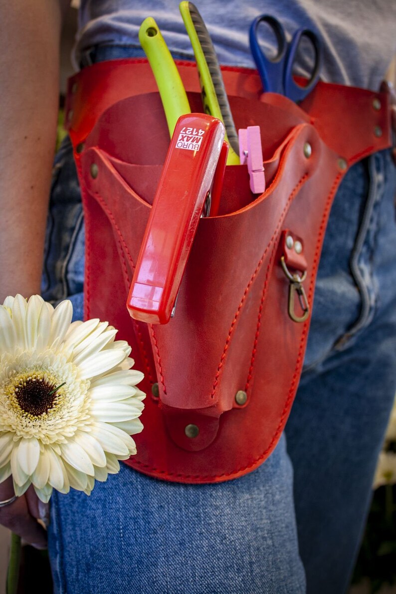 K&ouml;nnte beinhalten: Ein roter Leder-Werkzeugg&uuml;rtel mit einem roten Hefter, einer gr&uuml;nen und silbernen Schere und einem Paar rosa W&auml;scheklammern. Der G&uuml;rtel wird &uuml;ber einer blauen Jeans getragen.
