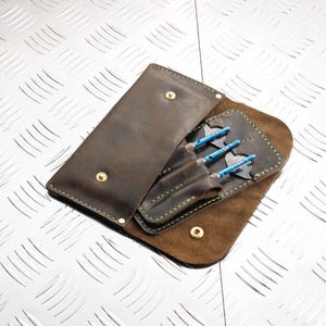 May include: Open leather dart case in shades of brown, with three blue and silver darts visible. The case is open, revealing the darts secured in individual slots. The case is secured with a gold snap closure and green stitching.