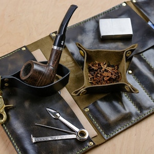 May include: A leather tobacco pipe roll with a dark brown pipe, a silver lighter, a small leather tray with tobacco, and a multi-tool. The roll is dark green with gold hardware and green stitching.