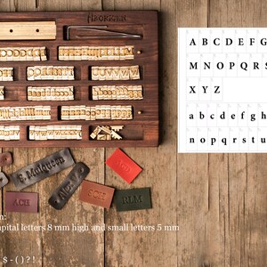 May include: A wooden box with a set of metal letter stamps for stamping leather. The box has a variety of letter stamps, including uppercase and lowercase letters, numbers, and symbols. The box also includes a small tool for pressing the stamps into the leather. The text on the box reads "BORGEN".