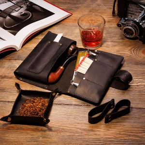 Puede incluir: Una bolsa de tabaco de cuero negro con una pipa, cerillas y una bandeja pequeña para tabaco. La bolsa tiene varios bolsillos y una correa.