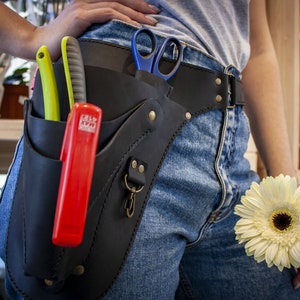 K&ouml;nnte beinhalten: Ein schwarzer Leder-Werkzeugg&uuml;rtel mit mehreren Taschen und einem Metallclip. Der G&uuml;rtel wird um die Taille getragen und enth&auml;lt eine Schere, ein rotes Universalmesser und ein gr&uuml;n-gelbes Universalmesser.