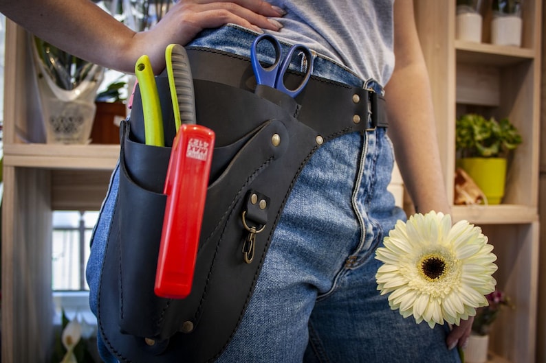 K&ouml;nnte beinhalten: Ein schwarzer Leder-Werkzeugg&uuml;rtel mit mehreren Taschen und einem Metallclip. Der G&uuml;rtel wird um die Taille getragen und enth&auml;lt eine Schere, ein rotes Universalmesser und ein gr&uuml;n-gelbes Universalmesser.