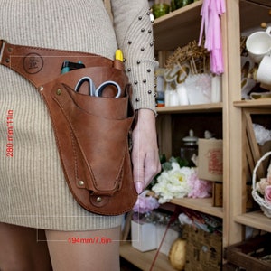 K&ouml;nnte beinhalten: Ein brauner Leder-Werkzeugg&uuml;rtel mit einer gro&szlig;en Tasche an der Seite. Der G&uuml;rtel ist 280 mm/11 Zoll lang und 194 mm/7,6 Zoll breit. Die Tasche ist mit Werkzeugen gef&uuml;llt, darunter eine Schere und ein Stift.