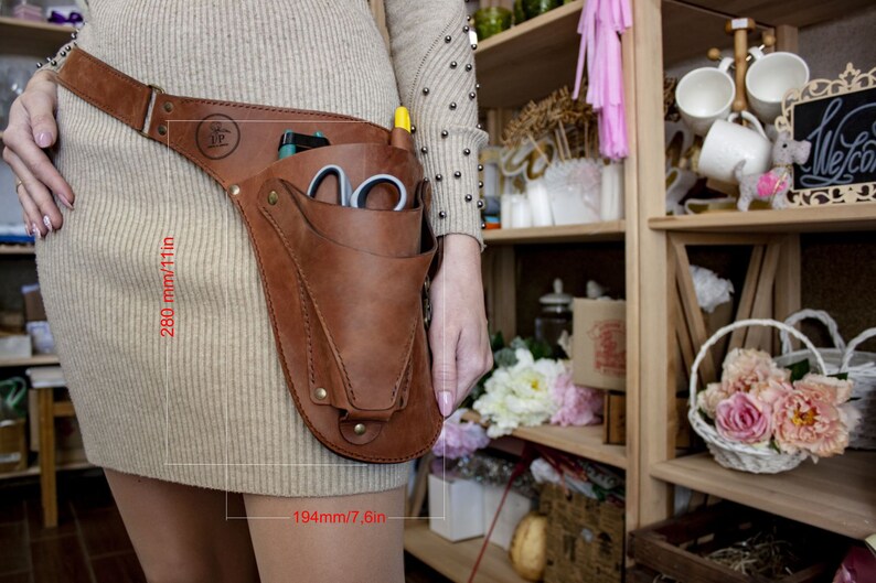 K&ouml;nnte beinhalten: Ein brauner Leder-Werkzeugg&uuml;rtel mit einer gro&szlig;en Tasche an der Seite. Der G&uuml;rtel ist 280 mm/11 Zoll lang und 194 mm/7,6 Zoll breit. Die Tasche ist mit Werkzeugen gef&uuml;llt, darunter eine Schere und ein Stift.
