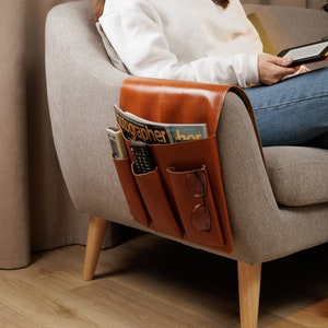 Könnte beinhalten: Ein brauner Leder-Armlehnen-Organizer mit mehreren Taschen zum Verstauen von Gegenständen wie Fernbedienungen, Zeitschriften und Brillen. Der Organizer ist an der Armlehne eines grauen Sofas befestigt.