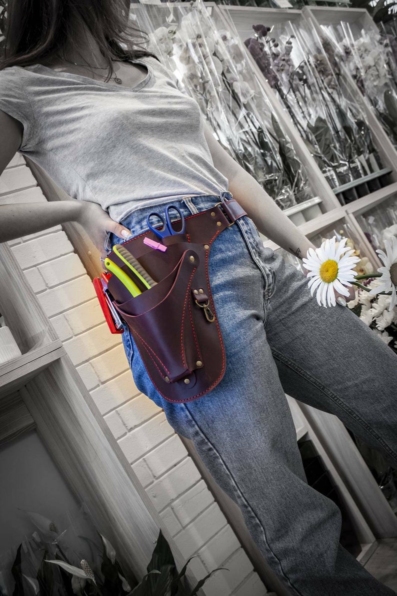 K&ouml;nnte beinhalten: Ein brauner Leder-Werkzeugg&uuml;rtel mit mehreren Taschen und roten N&auml;hten. Der G&uuml;rtel wird &uuml;ber einer blauen Jeans und einem grauen T-Shirt getragen. Der G&uuml;rtel enth&auml;lt verschiedene Werkzeuge, darunter eine Schere, einen Rasierer und einen Kamm.