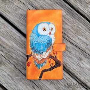 May include: Orange leather wallet with a detailed illustration of a blue and white owl on a branch with red berries. The wallet has a snap closure. The text "GALARY LEATHERCRAFT" is visible.