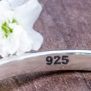 May include: Close-up of a silver band with the number "925" engraved on it. A white flower is in the background. The band is likely a piece of jewelry, possibly a ring or bracelet.