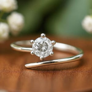May include: A silver ring with a large, clear, round gemstone at the center. The gemstone is held in place by six small prongs. The ring's band curves around the finger in a modern, minimalist design.