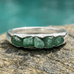Könnte beinhalten: Ein Silberring mit einer Reihe von fünf rohen, ungeschliffenen grünen Smaragden, die entlang der Vorderseite des Rings eingelassen sind. Das Metallband hat eine polierte, reflektierende Oberfläche, die mit den rauen Edelsteinen kontrastiert.