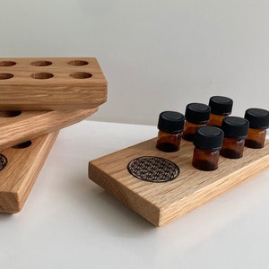 Puede incluir: Un juego de tres bandejas de madera con un diseño geométrico, apiladas una encima de la otra. La bandeja superior tiene ocho agujeros redondos para sostener botellas pequeñas. Una bandeja de madera separada contiene ocho pequeñas botellas de vidrio ámbar con tapas negras.