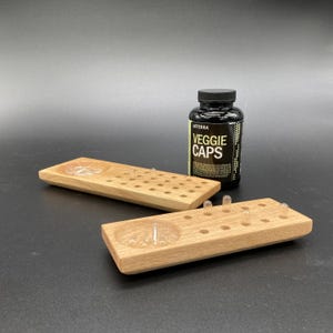 May include: Two wooden trays with clear capsules and a black bottle labelled "Veggie Caps". The trays have a rectangular shape with a rounded indentation and several small holes. The bottle is black with white text.