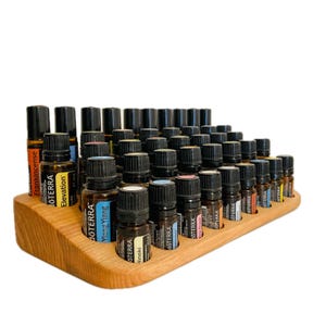 Op de afbeelding: Een houten displaystandaard voor essentiële oliën met talrijke kleine glazen flesjes met zwarte doppen. De flesjes zijn gelabeld met verschillende namen van essentiële oliën en het merk "doTERRA". De standaard is gemaakt van lichtgekleurd hout.