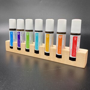 May include: A wooden essential oil holder with seven roller bottles. Each bottle has a white cap and a label with a different color and name: Rescuer, Calmer, Steady, Tamer, Thinker, Brave, and Stronger. The holder is made of light-colored wood.
