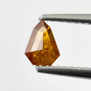 May include: A faceted, amber-coloured gemstone held by metal tweezers. The gem has a unique, geometric shape with multiple facets that reflect light. The colour ranges from a deep orange to a lighter, golden hue, showcasing its clarity and brilliance.