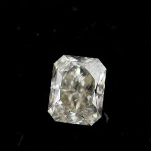 May include: A radiant-cut gemstone with a clear, translucent appearance and a multifaceted surface that reflects light. The stone features a rectangular shape with clipped corners, displaying internal light refractions and a polished finish.