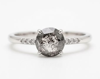 May include: A silver-toned ring featuring a large, round, salt-and-pepper diamond set in a four-prong setting. The band is accented with three small, clear, round-cut diamonds on each side of the central stone, creating a balanced look.
