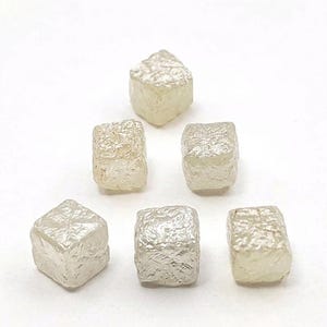 Könnte beinhalten: Sechs roh geschnittene, würfelförmige Diamanten sind vor einem weißen Hintergrund angeordnet. Die Diamanten haben einen blassgelben Farbton und eine strukturierte Oberfläche, die das Licht reflektiert. Die Edelsteine sind unterschiedlich groß und zeigen ihren natürlichen, unpolierten Zustand.