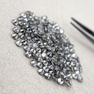 May include: A collection of numerous small, faceted, dark gray salt and pepper diamonds arranged in a cluster. Each stone features a brilliant cut with multiple reflective surfaces, displaying a metallic luster and a translucent, speckled appearance.
