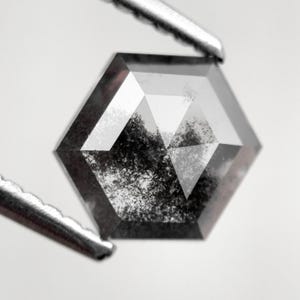 May include: A hexagonal-cut, salt and pepper diamond held by a pair of silver tweezers. The gemstone has a dark gray color with internal inclusions, and facets that reflect light. The diamond is set against a white background.
