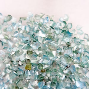 Peut inclure: Une collection de petits éclats de verre translucides et irréguliers dans des tons bleu clair, sarcelle et vert pâle. Les fragments présentent des bords tranchants et anguleux ainsi qu'une surface brillante et réfléchissante.