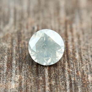 May include: A round, faceted, translucent gemstone with a pale, icy gray-white color. The stone features a brilliant cut with numerous triangular and kite-shaped facets that reflect light, creating a sparkling appearance across its circular surface.