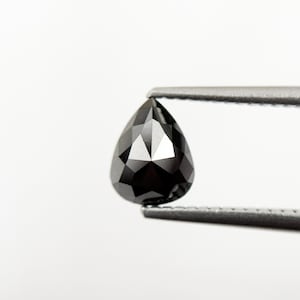 May include: A faceted, pear-shaped black diamond held by silver tweezers against a white background. The diamond's surface reflects light, showcasing its many facets and dark color. The gemstone is a striking example of fine jewelry.
