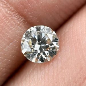 Puede incluir: Una piedra preciosa de corte brillante redondo con una apariencia clara e incolora y múltiples facetas triangulares y en forma de cometa que reflejan la luz. La piedra presenta un contorno circular simétrico y una estructura interna compleja de superficies geométricas.