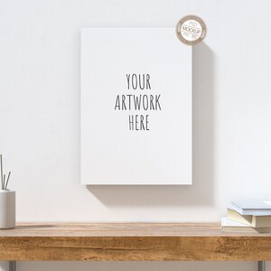 May include: A white framed canvas mockup with the text "YOUR ARTWORK HERE" on a wooden table with a white vase and a stack of books.