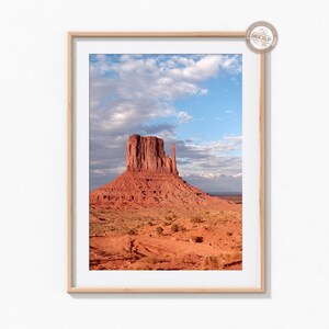 May include: A framed photograph of a red rock formation in a desert landscape. The sky is blue with white clouds. The rock formation is tall and thin, with a flat top. The landscape is mostly red sand and dirt, with some green vegetation.