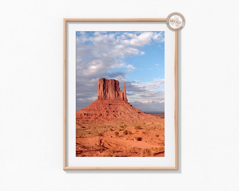 May include: A framed photograph of a red rock formation in a desert landscape. The sky is blue with white clouds. The rock formation is tall and thin, with a flat top. The landscape is mostly red sand and dirt, with some green vegetation.