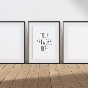 Puede incluir: Tres marcos de fotos negros con paspartú blanco colgados en una pared blanca. El marco del medio tiene el texto "YOUR ARTWORK HERE" en negro.