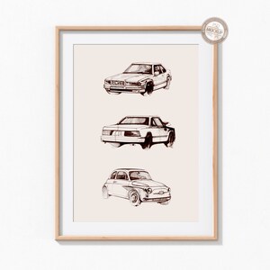 May include: A framed print of three vintage cars drawn in black and white ink. The cars are a sedan, a hatchback, and a small car. The print is on a light beige background.