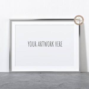 Könnte beinhalten: Ein rechteckiger silberner Bilderrahmen mit weißem Passepartout, der den Text "YOUR ARTWORK HERE" anzeigt. Der Rahmen steht auf einer grauen Oberfläche vor einer weißen Wand. Ein kleiner, runder Aufkleber befindet sich in der oberen rechten Ecke.