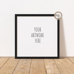 May include: A square black picture frame with the text "YOUR ARTWORK HERE" on a white background. The frame is leaning against a white wall on a wooden floor. A small circular sticker is in the upper right corner.